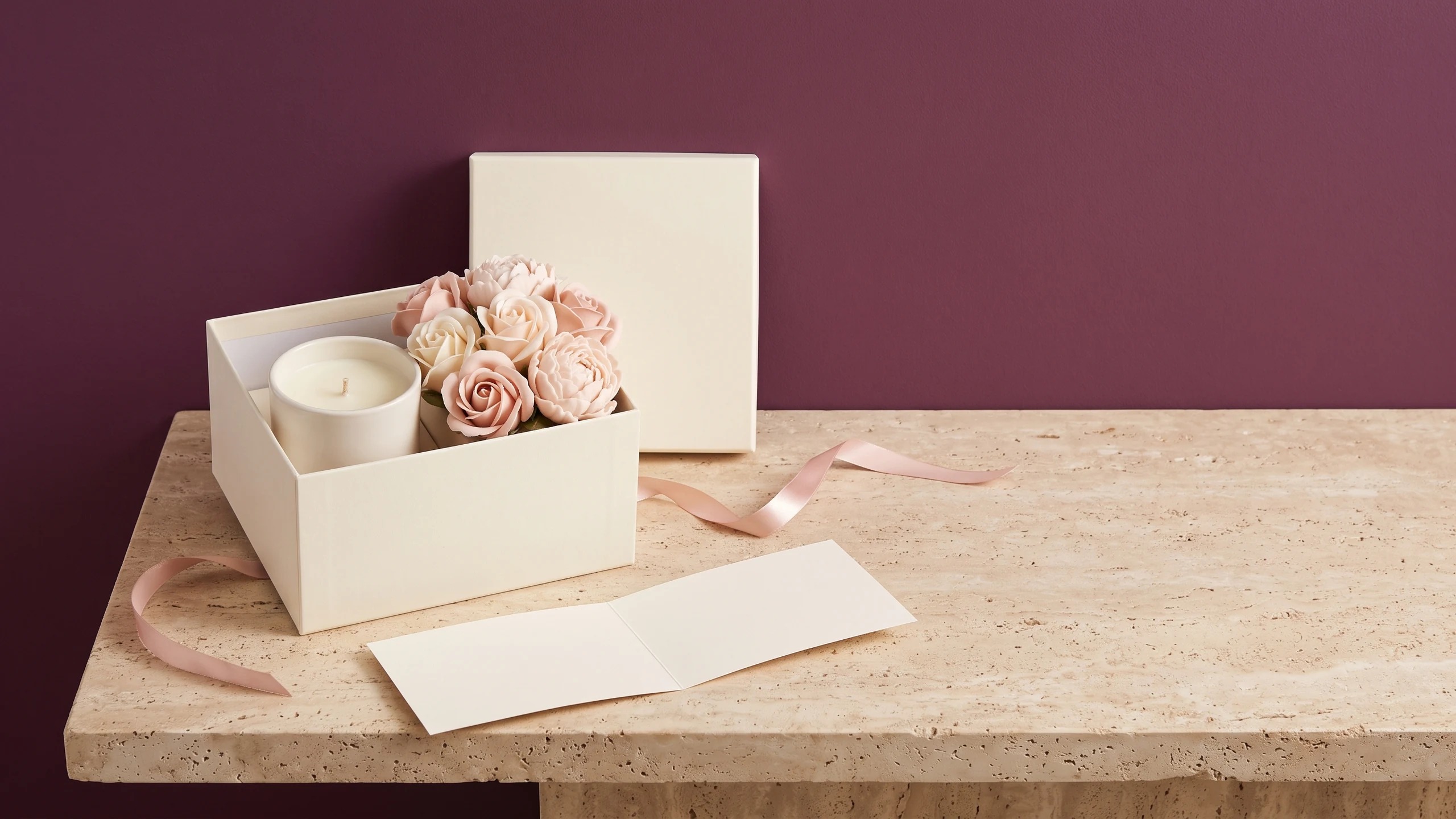 An open cream gift box holding a candle and blush soap roses, with the lid behind, untied pink ribbon and a blank open card, against a plum wall