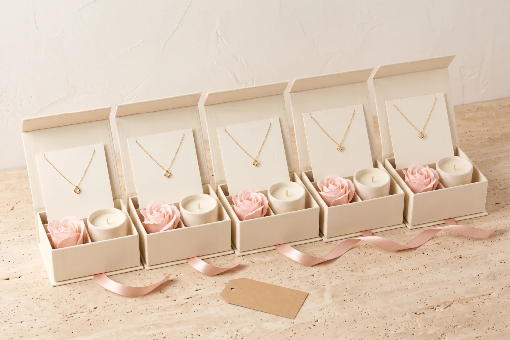 Five open cream gift boxes in a row, each holding a gold pendant necklace, a pink soap rose and a cream candle, with pink ribbon and a kraft tag