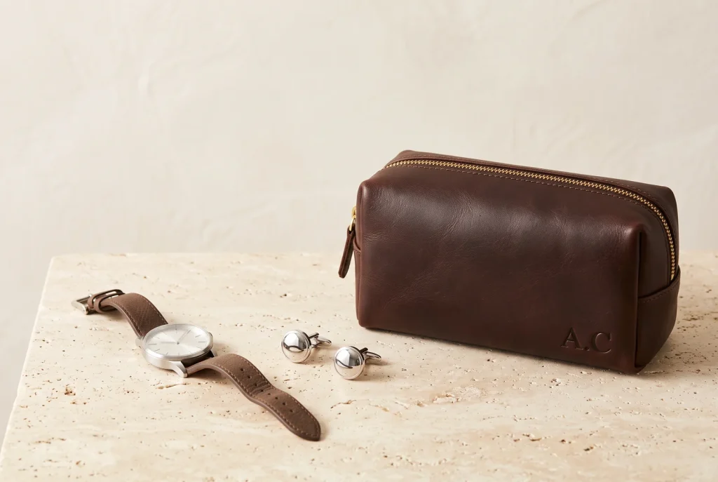 A brown leather dopp kit monogrammed with two initials on travertine, with polished silver cufflinks and a men's watch on a brown leather strap beside it