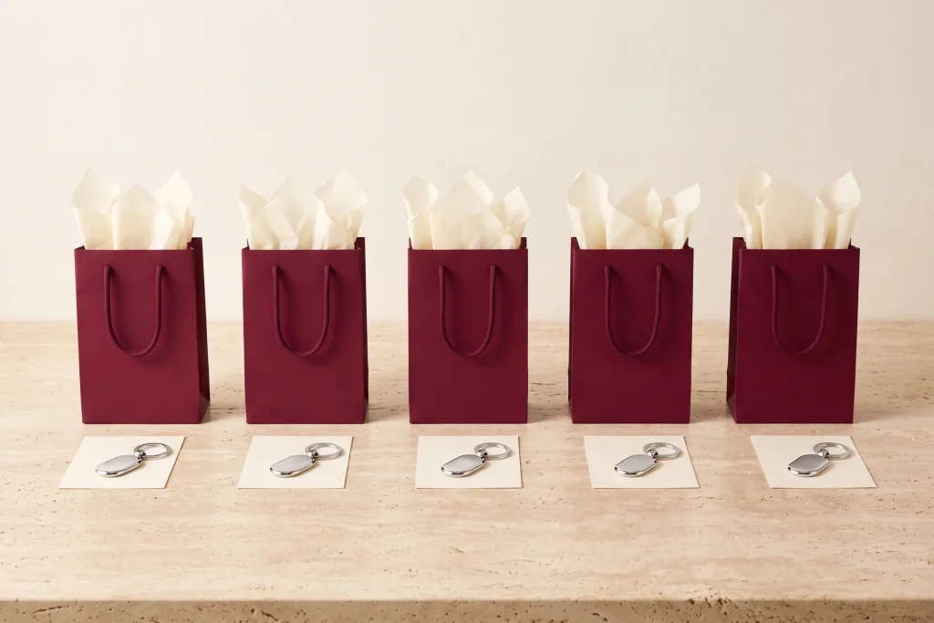 Five matching burgundy gift bags with cream tissue in a row, each with a blank card and a silver keychain set out in front, on travertine