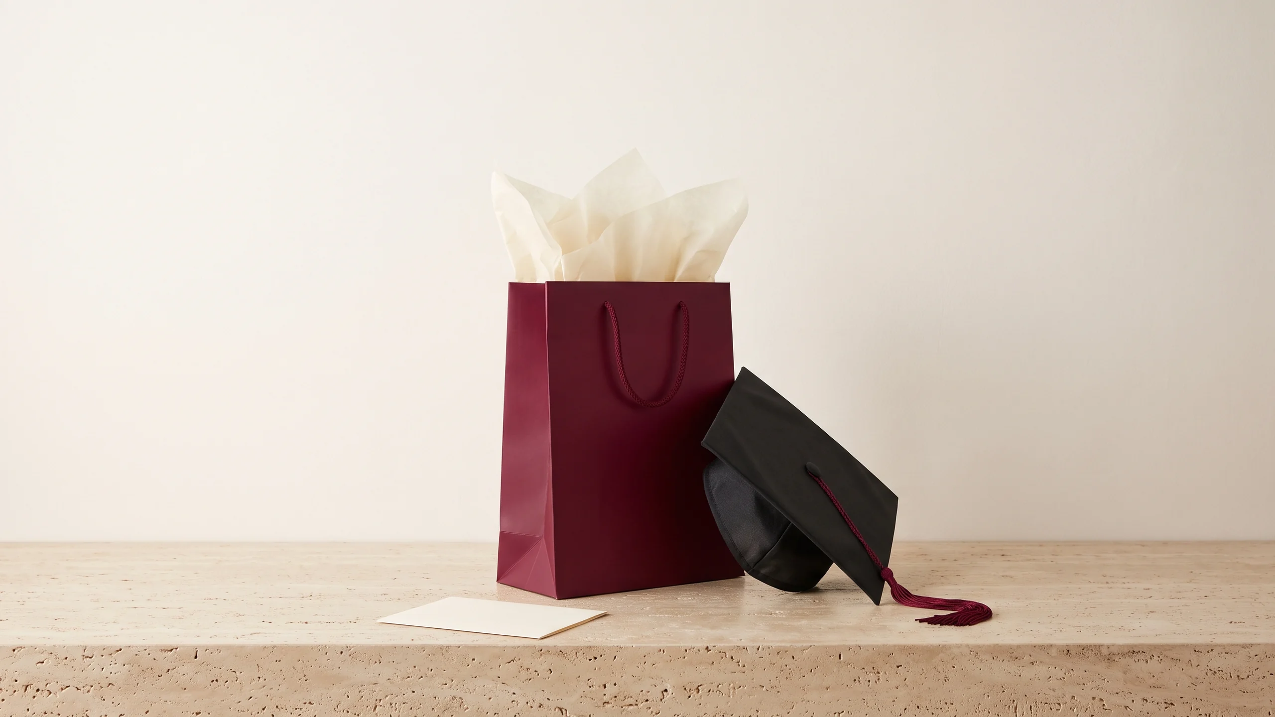 Deep burgundy gift bag with cream tissue paper, a black graduation cap leaning against it and a blank card in front, on travertine