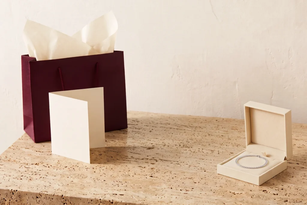 Burgundy gift bag with cream tissue beside an open blank card and an open jewelry box holding a silver chain bracelet, on travertine