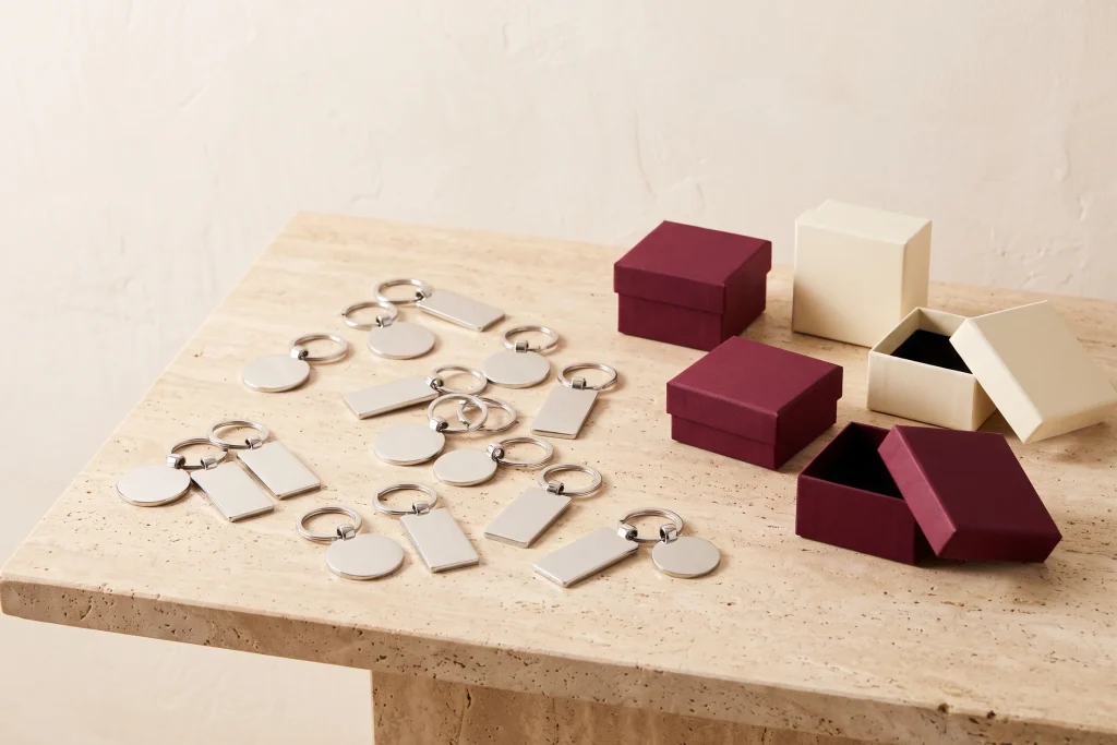 Polished silver keychains with blank tags scattered on travertine beside small cream and burgundy gift boxes, sorted for a class order