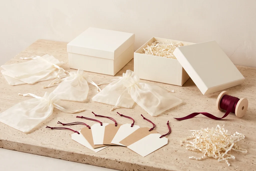 Ivory gift boxes, sheer organza bags, blank kraft tags on burgundy cord and a spool of burgundy ribbon laid out for assembly
