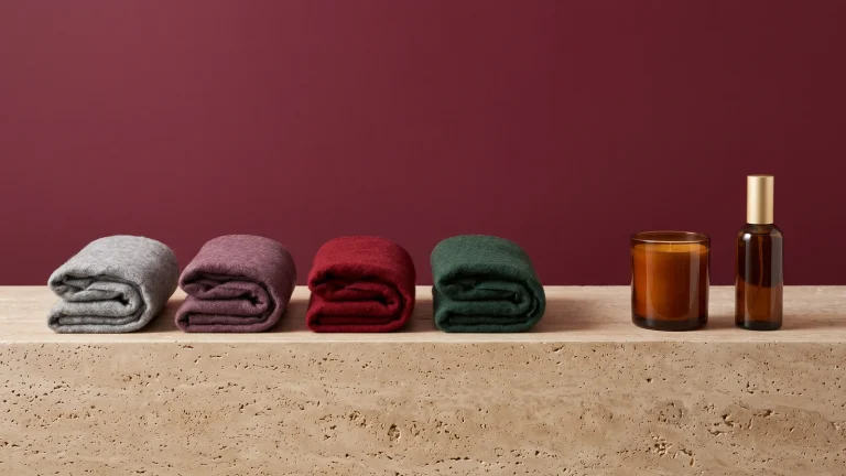 Folded wool scarves in grey, mauve, red and green beside an amber candle and an amber room spray with a gold cap on travertine against a burgundy wall