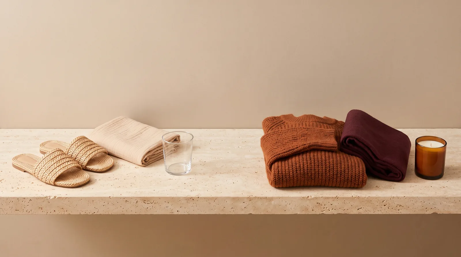 Raffia sandals, a folded sand cloth and a glass tumbler beside a rust knit sweater, a burgundy fold and an amber candle on travertine
