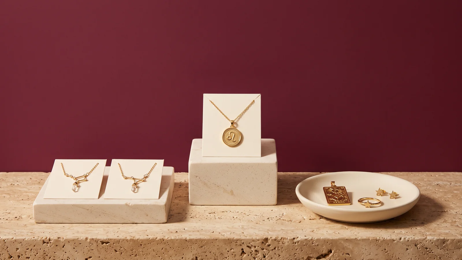 Gold constellation necklaces and a Leo coin pendant on cream cards and stone risers, with a tarot pendant, star ring and star earrings in a dish, against a burgundy wall