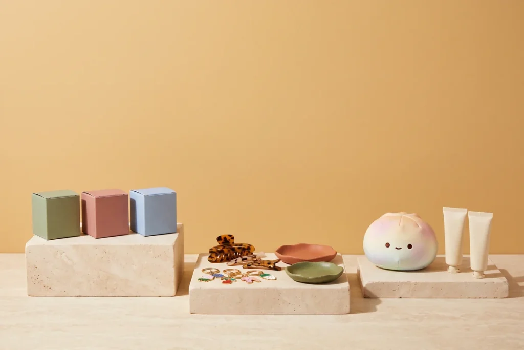 Small blank boxes, enamel charm keychains, hair clips, trinket dishes, a pastel plush and hand cream tubes arranged on three stone risers against an ochre wall