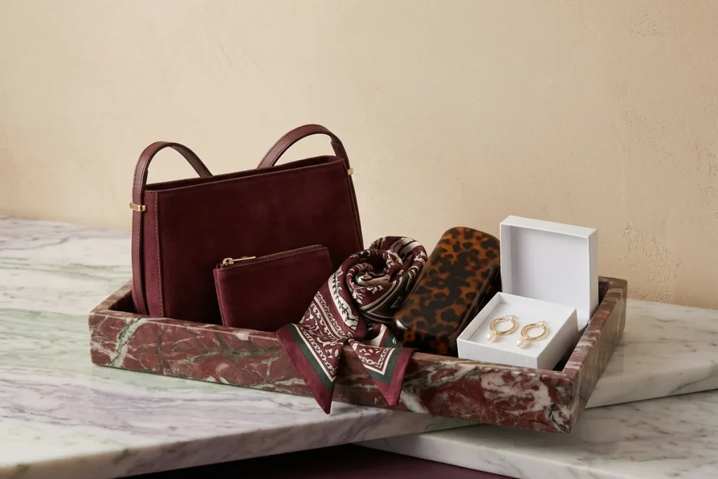 Rows of matching gold hoop earrings in open white boxes, stacks of closed white boxes behind them and a linen tray holding more pairs on a black and gold marble shelf against a lilac wall