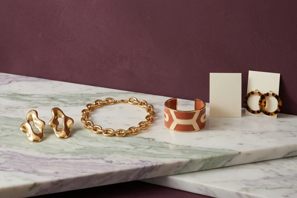 Sculptural gold earrings, a chunky gold chain bracelet, a terracotta and cream enamel cuff and tortoiseshell resin hoops on a cream card on a white marble slab with green and lilac veining against a plum wall