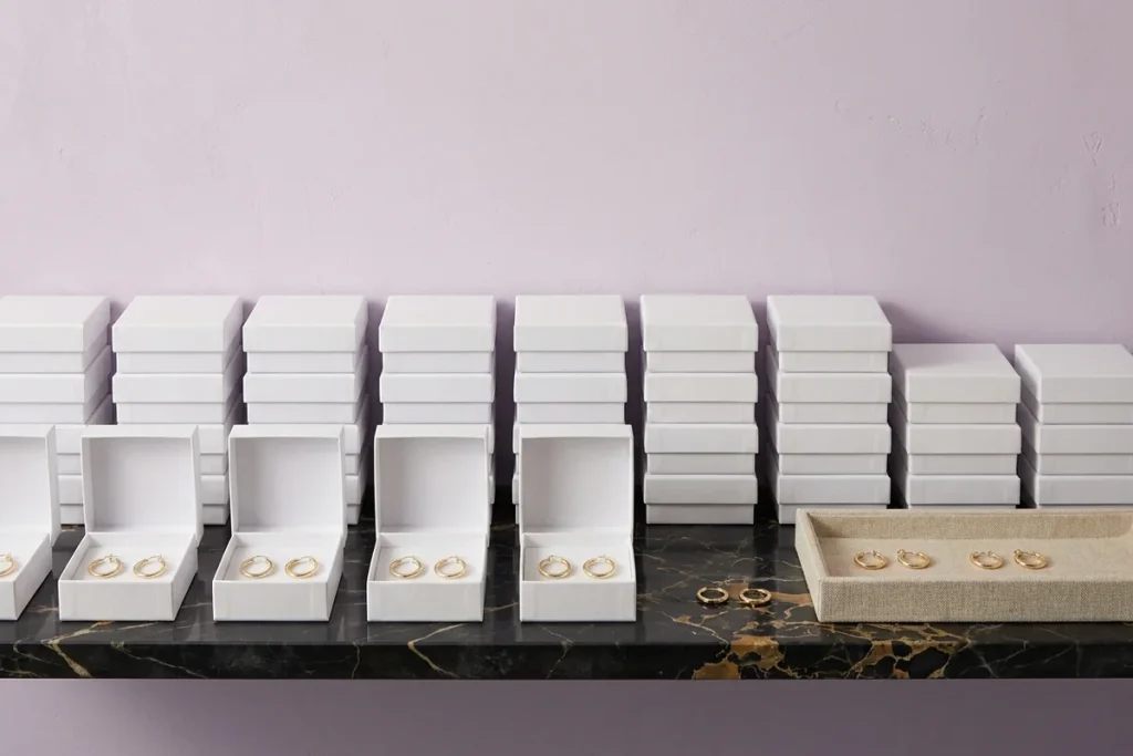 Rows of matching gold hoop earrings in open white boxes, stacks of closed white boxes behind them and a linen tray holding more pairs on a black and gold marble shelf against a lilac wall