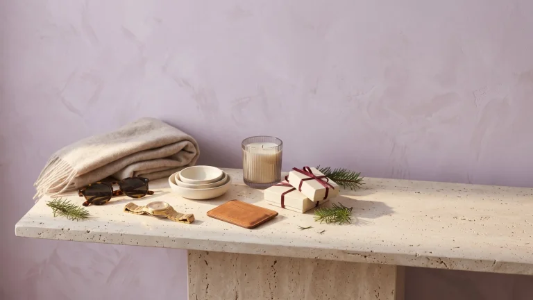 A folded cashmere scarf, tortoiseshell sunglasses, a gold watch, cream serving dishes, a leather card wallet, a ribbed candle and two gift boxes tied with burgundy ribbon on a travertine console against a lilac wall