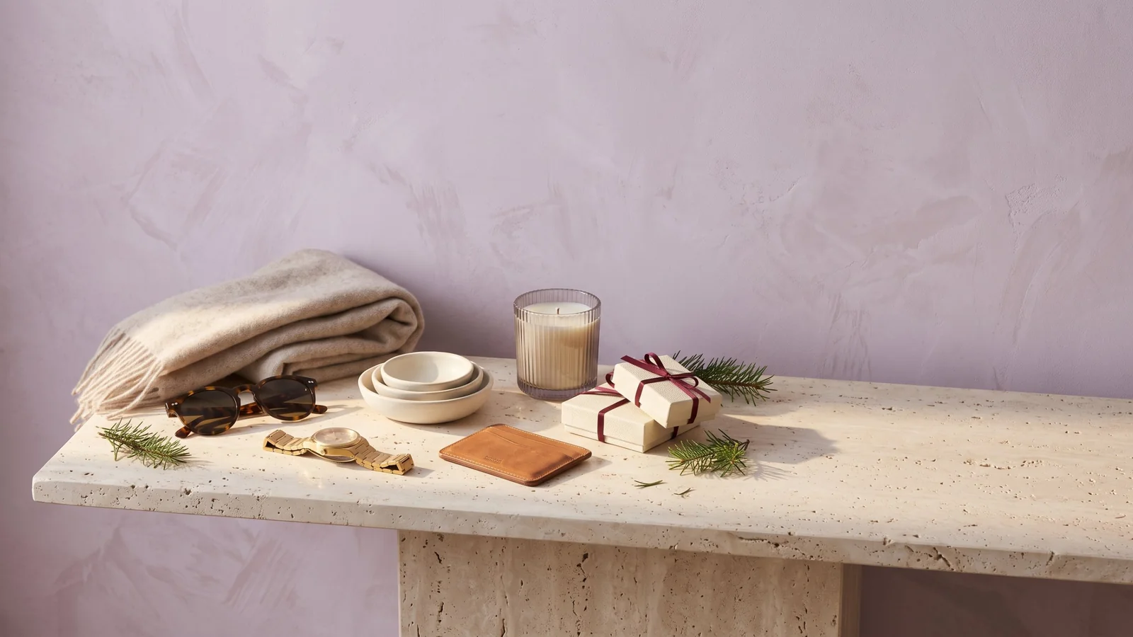 A folded cashmere scarf, tortoiseshell sunglasses, a gold watch, cream serving dishes, a leather card wallet, a ribbed candle and two gift boxes tied with burgundy ribbon on a travertine console against a lilac wall
