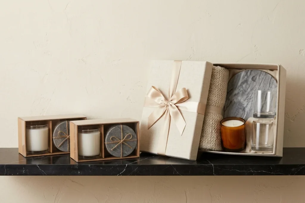 Two kraft gift boxes with clear windows, each holding a white candle and twine-tied stone coasters, beside a linen gift box with a satin bow opened to show a knit throw, an amber candle, a grey marble board and two glass tumblers on a black marble shelf