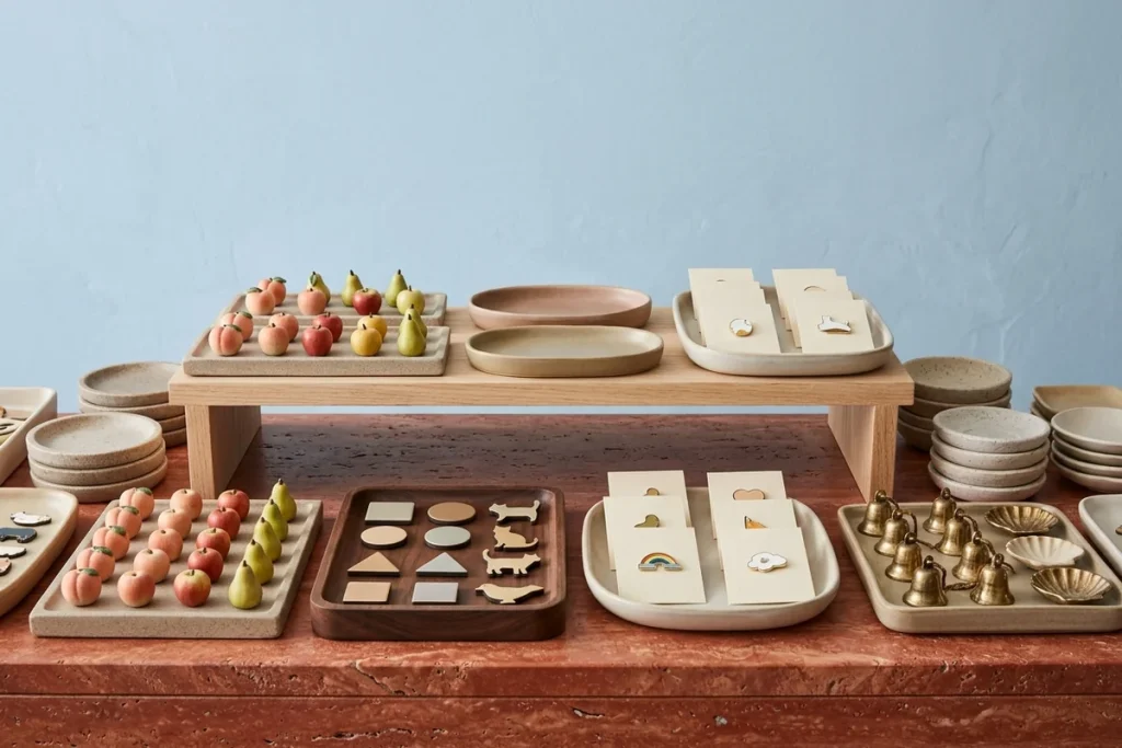 A red travertine counter with an oak riser against a sky-blue wall, holding trays of ceramic peaches, apples and pears, geometric and animal magnets, enamel pins on blank cards, brass bells, shell dishes and stacked speckled trinket dishes
