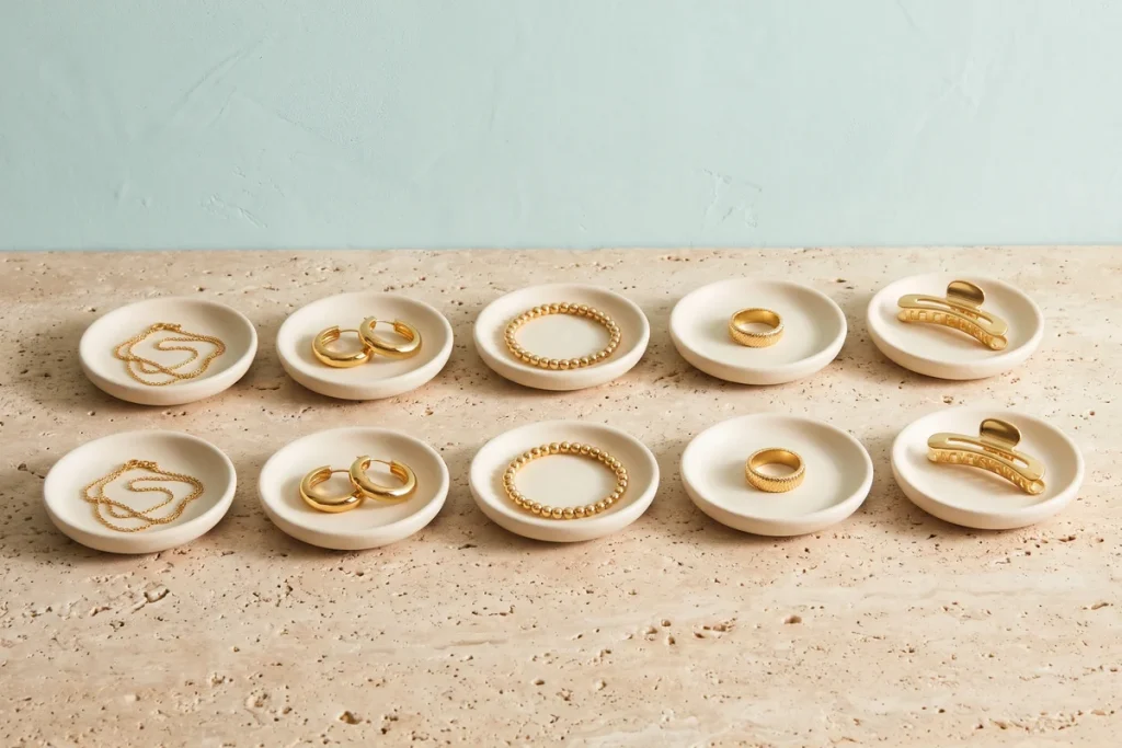 Two matching rows of gold jewelry and hair clips on cream ceramic dishes, each chain, hoop, bracelet, ring and clip identical to the one behind it