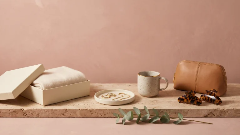 A cashmere scarf in an open gift box, gold hoops and a chain on a ceramic tray, a gold-rimmed mug, a tan leather pouch and claw clips on a stone ledge against a dusty rose wall