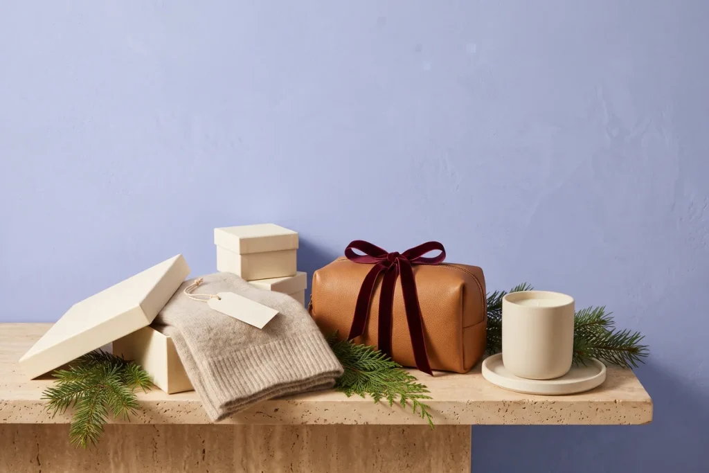 A tan leather travel pouch tied with burgundy velvet ribbon, a knit scarf in an open gift box and a candle on a tray, surrounded by pine sprigs against a periwinkle wall