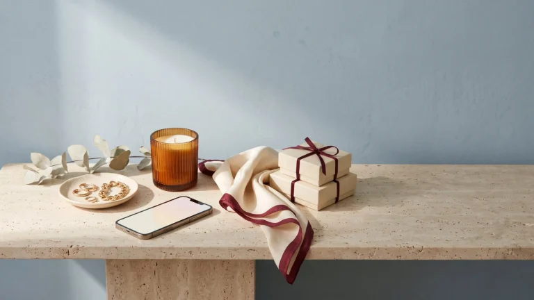 A smartphone beside gold hoop earrings and a gold chain bracelet on a cream dish, an amber candle, a folded silk scarf and two gift boxes tied with burgundy ribbon on a travertine console against a powder-blue wall