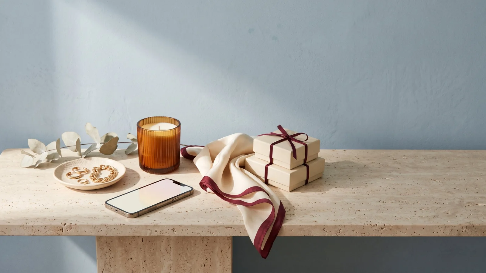 A smartphone beside gold hoop earrings and a gold chain bracelet on a cream dish, an amber candle, a folded silk scarf and two gift boxes tied with burgundy ribbon on a travertine console against a powder-blue wall