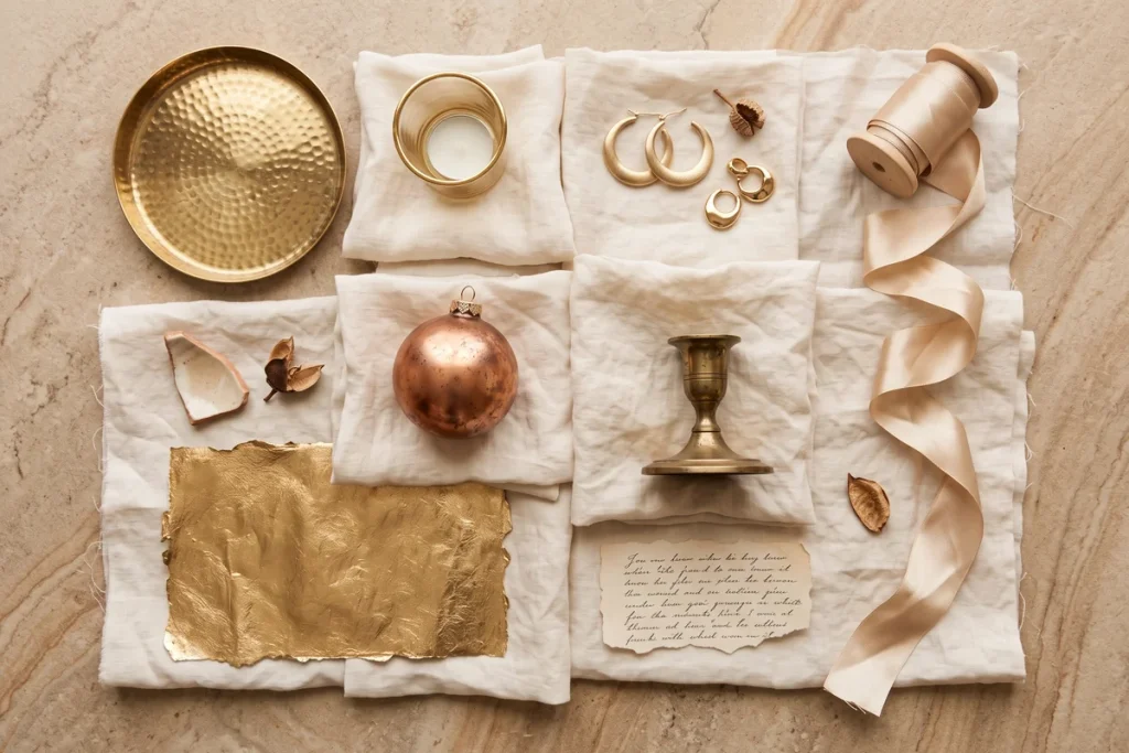 An overhead mood-board flat lay of warm metallic pieces, including a hammered brass tray, gold hoop earrings, a gold-rimmed votive, a copper ornament and champagne ribbon on stone