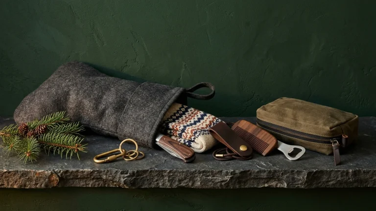 A grey felt stocking on a rough slate ledge against a forest green wall, spilling fair isle wool socks, a wooden-handled pocket knife, a brass carabiner keyring, a leather cord organizer, a wooden comb, a bottle opener and a waxed canvas pouch beside a pine sprig