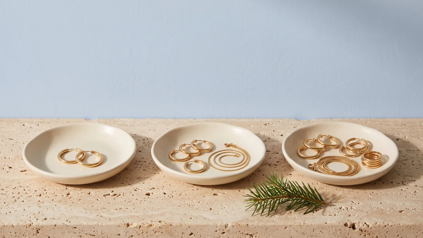 Three cream ceramic dishes of matching gold hoops, chains and rings, filling from left to right against a powder blue wall