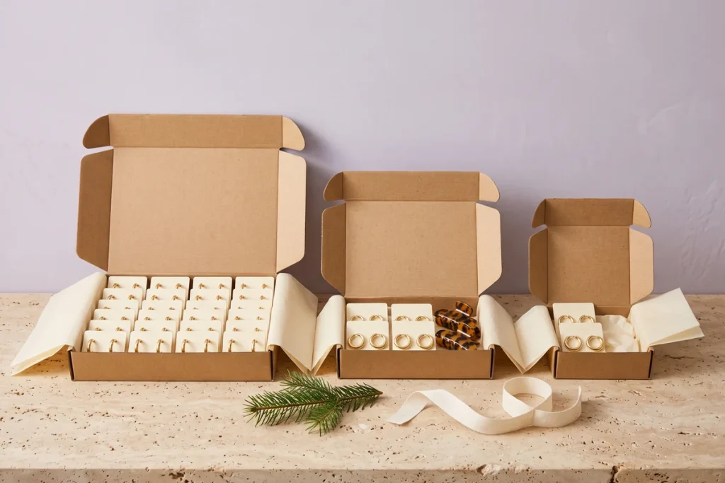 Three open kraft boxes of decreasing size holding gold hoop earrings and claw clips, with a pine sprig and cream ribbon beside them against a lilac grey wall
