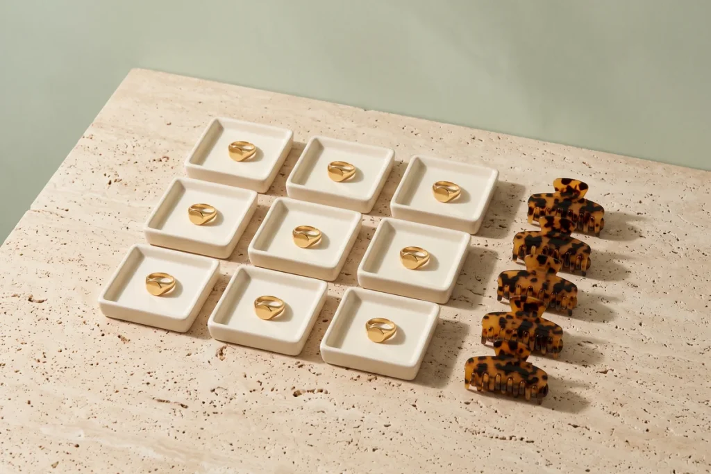 A grid of nine cream trays each holding an identical gold signet ring, beside a row of five matching tortoiseshell claw clips against a sage green wall