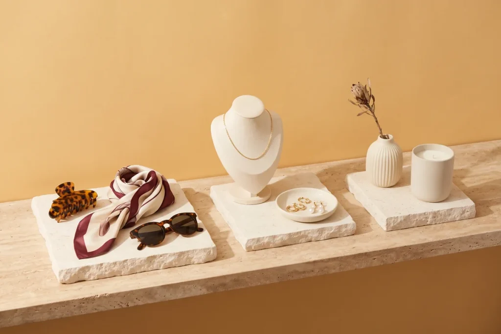 Claw clips, a silk scarf and sunglasses, then a gold necklace and earrings, then a ceramic vase and candle on three separate stone risers against a marigold wall