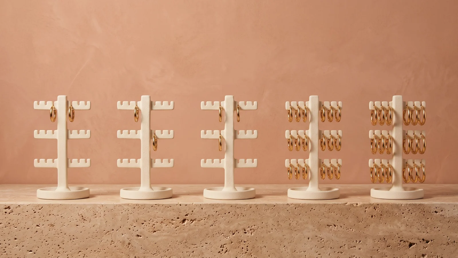 Five cream jewelry stands on a stone ledge, three nearly empty with a few gold hoops left and two fully stocked, against a terracotta wall
