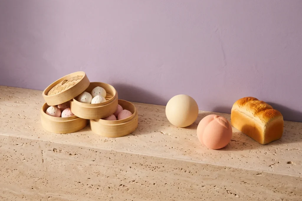 Dumpling squishies in stacked bamboo steamers beside a cream stress ball, a peach squeeze toy and a bread-loaf squishy against a lilac wall