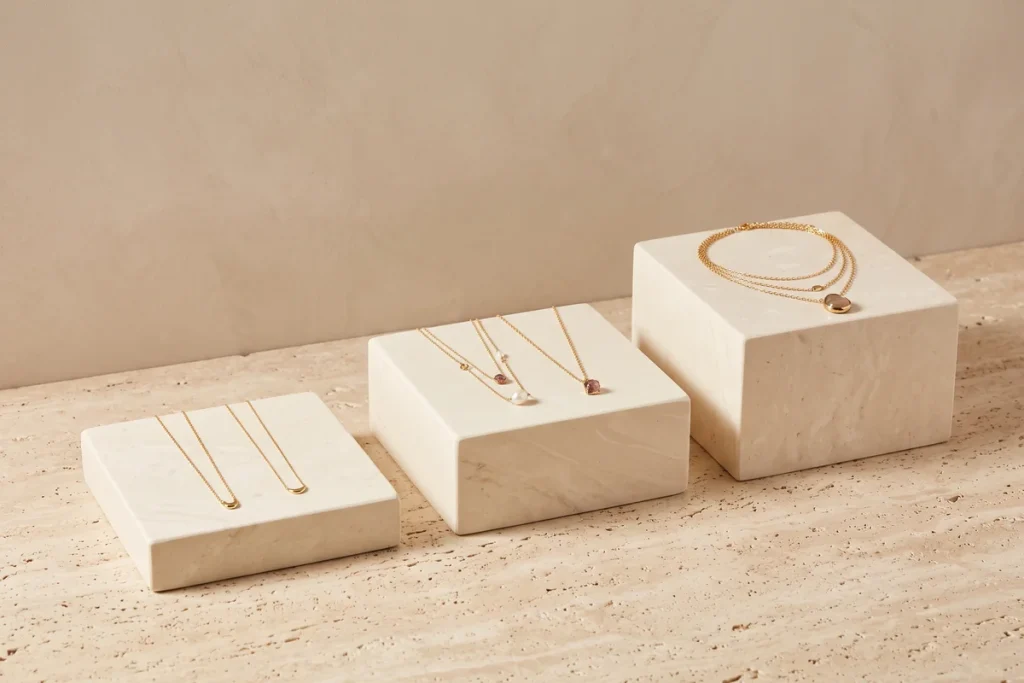 Gold necklaces on three cream stone risers of stepped heights, from two fine chains on the lowest to pearl and stone pendants in the middle and a layered necklace on the tallest