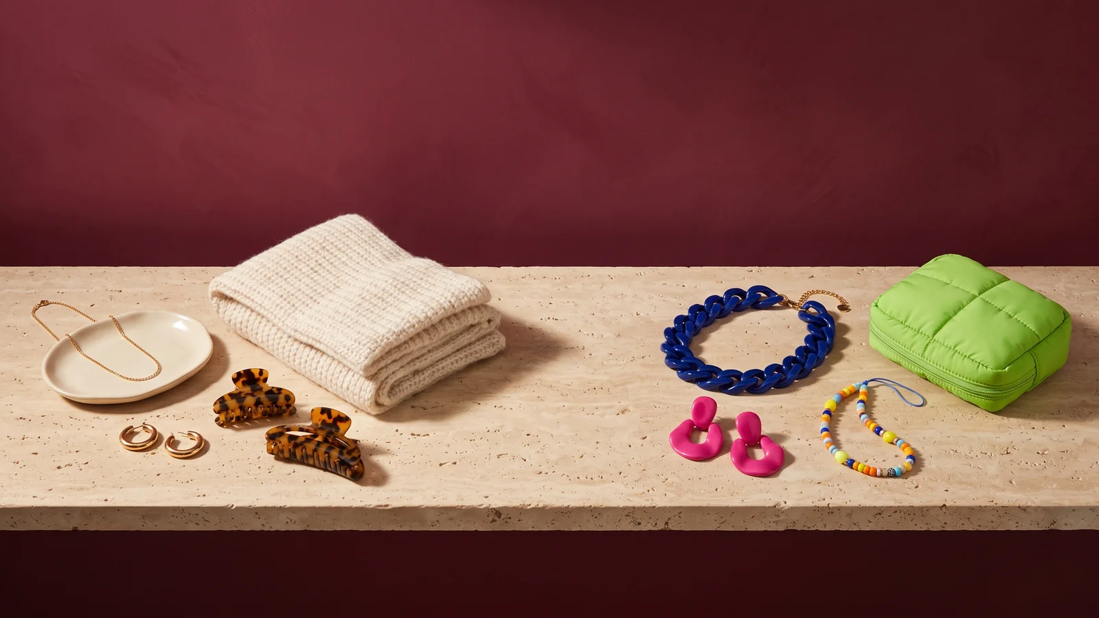 A gold chain on a cream dish, gold hoops, tortoiseshell claw clips and a cream knit scarf beside a cobalt chain necklace, pink earrings, a beaded phone strap and a lime quilted pouch on travertine against a burgundy wall