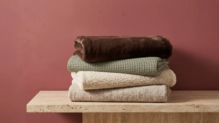 A stack of folded oatmeal fleece, cream sherpa, sage waffle-knit and brown faux mink throws on a stone ledge against a dusty cranberry wall