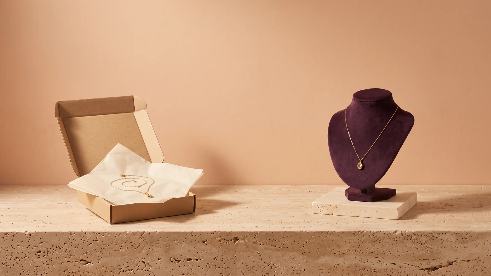 A gold pendant necklace coiled on tissue in a plain kraft mailer box beside the same necklace displayed on a plum velvet bust, on travertine against a peach wall
